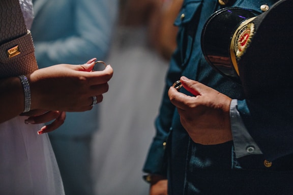 Two individuals facing each other exchange rings. One person wears a formal military uniform complete with a decorated hat under their arm, while the other is dressed in a gown with jewelry. The setting is dimly lit, suggesting a formal or ceremonial atmosphere.