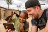 A young boy and a man share a joyful moment while looking at a smartphone. The boy is smiling broadly and wearing a patterned shirt, while the man, wearing a cap and a backpack, looks delighted. They are seated outdoors near a rustic, mud-brick structure with a tree and cloudy sky in the background, suggesting a rural setting.