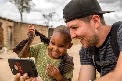 A young boy and a man share a joyful moment while looking at a smartphone. The boy is smiling broadly and wearing a patterned shirt, while the man, wearing a cap and a backpack, looks delighted. They are seated outdoors near a rustic, mud-brick structure with a tree and cloudy sky in the background, suggesting a rural setting.