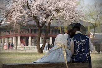 A couple dressed in traditional Korean hanbok sit closely on a bench, with the woman resting her head on the man's shoulder. In the background, cherry blossom trees are in full bloom, and a historical building with columns and red accents is visible. Other people can be seen walking and also dressed in traditional clothing, enhancing the cultural ambiance.