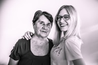 An older woman with short hair stands beside a younger woman with long blonde hair and glasses. They both have relaxed and happy expressions, with one arm draped affectionately across the older woman's shoulder.