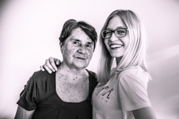 An older woman with short hair stands beside a younger woman with long blonde hair and glasses. They both have relaxed and happy expressions, with one arm draped affectionately across the older woman's shoulder.
