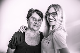 An older woman with short hair stands beside a younger woman with long blonde hair and glasses. They both have relaxed and happy expressions, with one arm draped affectionately across the older woman's shoulder.