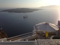 A serene ocean view with a cruise ship sailing near a small island. The foreground shows a balcony with white architectural elements, a large umbrella, and a seating area. The early morning or late afternoon sun casts a warm glow over the scene.