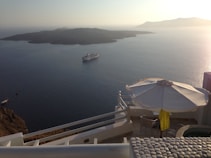 A serene ocean view with a cruise ship sailing near a small island. The foreground shows a balcony with white architectural elements, a large umbrella, and a seating area. The early morning or late afternoon sun casts a warm glow over the scene.