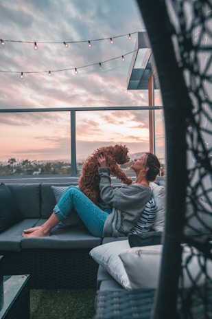 A person is sitting on an outdoor couch, holding a dog close to their face. The setting is a cozy balcony or patio during sunset, with a string of lights hanging above. The person is wearing casual clothing and appears relaxed and happy.