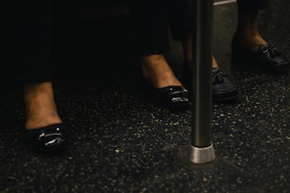 Feet wearing black shoes are positioned on a speckled floor, with a metal pole partially in view.
