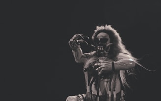 A figure in a theatrical, detailed costume that includes a mask with exaggerated features and long hair, performing a dramatic pose in front of a dark background.