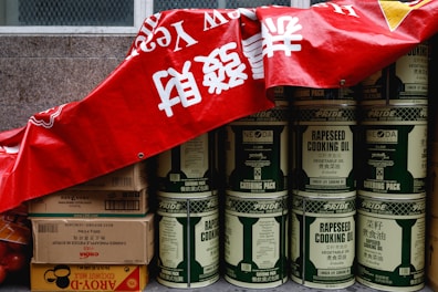 A stack of large metal cans labeled 'rapeseed cooking oil' is partially covered by a red banner with white characters. Beside the cans, there are several cardboard boxes, some labeled with brands and product information, including soy sauce and coconut milk. The cans and boxes are arranged against a stone wall, giving the impression of storage or preparation for a market or event.