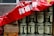 A stack of large metal cans labeled 'rapeseed cooking oil' is partially covered by a red banner with white characters. Beside the cans, there are several cardboard boxes, some labeled with brands and product information, including soy sauce and coconut milk. The cans and boxes are arranged against a stone wall, giving the impression of storage or preparation for a market or event.
