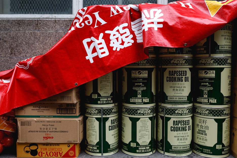 A stack of large metal cans labeled 'rapeseed cooking oil' is partially covered by a red banner with white characters. Beside the cans, there are several cardboard boxes, some labeled with brands and product information, including soy sauce and coconut milk. The cans and boxes are arranged against a stone wall, giving the impression of storage or preparation for a market or event.