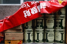 A stack of large metal cans labeled 'rapeseed cooking oil' is partially covered by a red banner with white characters. Beside the cans, there are several cardboard boxes, some labeled with brands and product information, including soy sauce and coconut milk. The cans and boxes are arranged against a stone wall, giving the impression of storage or preparation for a market or event.