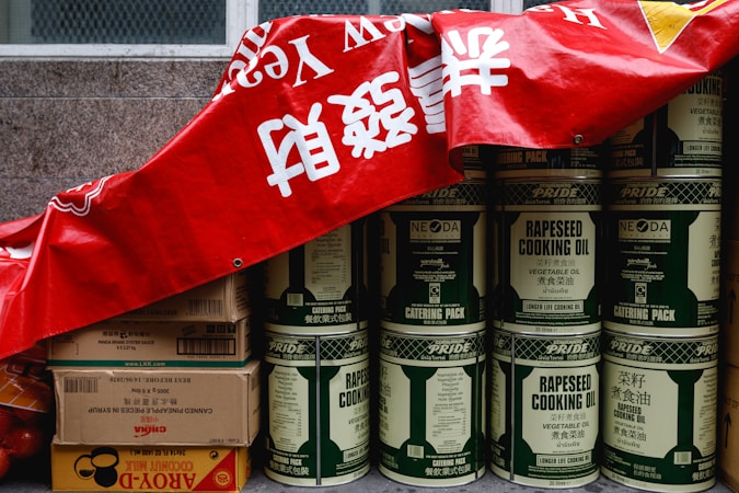 A stack of large metal cans labeled 'rapeseed cooking oil' is partially covered by a red banner with white characters. Beside the cans, there are several cardboard boxes, some labeled with brands and product information, including soy sauce and coconut milk. The cans and boxes are arranged against a stone wall, giving the impression of storage or preparation for a market or event.