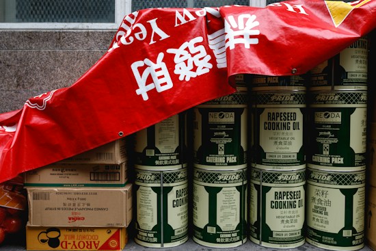 A stack of large metal cans labeled 'rapeseed cooking oil' is partially covered by a red banner with white characters. Beside the cans, there are several cardboard boxes, some labeled with brands and product information, including soy sauce and coconut milk. The cans and boxes are arranged against a stone wall, giving the impression of storage or preparation for a market or event.