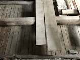 A detailed view of a wooden floor structure with overlapping planks and visible beams. The boards and beams appear weathered and aged, possibly part of a construction or renovation project. The surface shows signs of wear and some dust accumulation.