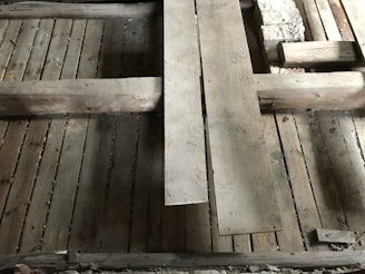 A detailed view of a wooden floor structure with overlapping planks and visible beams. The boards and beams appear weathered and aged, possibly part of a construction or renovation project. The surface shows signs of wear and some dust accumulation.