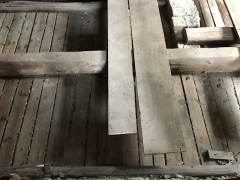 A detailed view of a wooden floor structure with overlapping planks and visible beams. The boards and beams appear weathered and aged, possibly part of a construction or renovation project. The surface shows signs of wear and some dust accumulation.