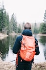 man wearing blue jacket and red backpack facing body of water