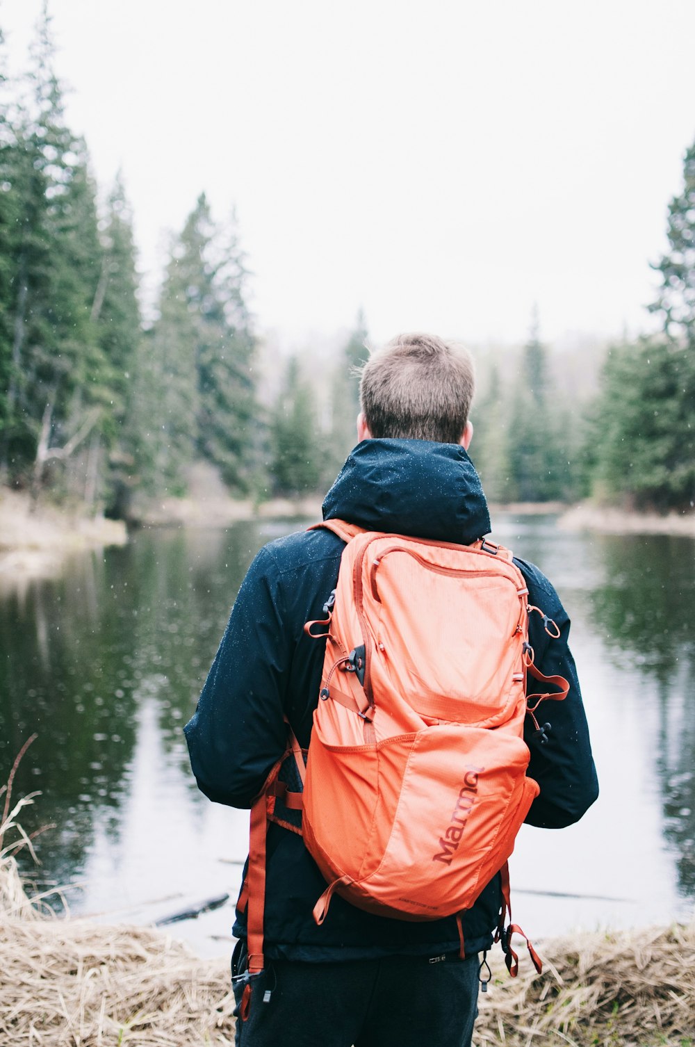 man wearing blue jacket and red backpack facing body of water