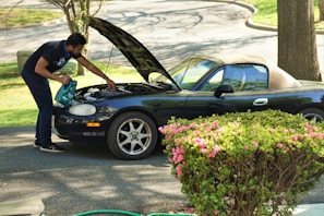 A person is inspecting the engine of a dark convertible car with the hood open. They are standing on a paved driveway surrounded by green bushes with pink flowers. The background includes trees and grassy areas.