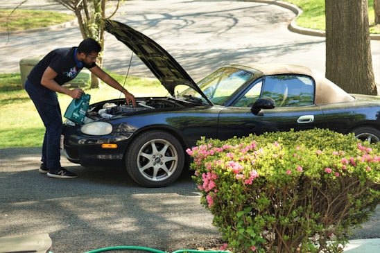 A person is inspecting the engine of a dark convertible car with the hood open. They are standing on a paved driveway surrounded by green bushes with pink flowers. The background includes trees and grassy areas.
