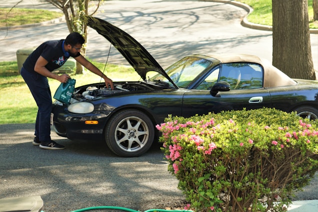 A person is inspecting the engine of a dark convertible car with the hood open. They are standing on a paved driveway surrounded by green bushes with pink flowers. The background includes trees and grassy areas.