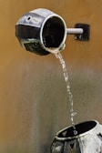 Water flows from a metal spout into a decorative metal container below. The spout is attached to a wall, and the entire setup creates a small outdoor water feature. The background wall has a weathered texture with a muted color tone.