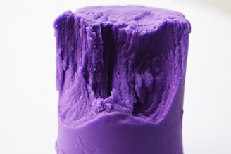 A close-up of a cylindrical piece of purple modeling clay with a rugged and indented texture on top. The clay appears soft and pliable.