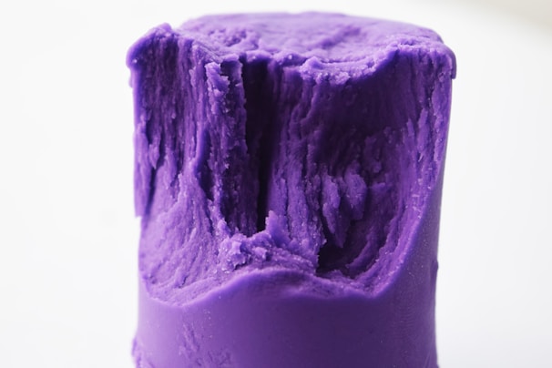 A close-up of a cylindrical piece of purple modeling clay with a rugged and indented texture on top. The clay appears soft and pliable.