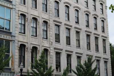 A multi-story, historic stone building with tall, narrow windows and distinctive architectural details. The facade shows signs of aging with some discoloration and weathering, accentuating its vintage charm. Several trees with lush green foliage are visible at street level, adding a touch of nature to the urban scene.