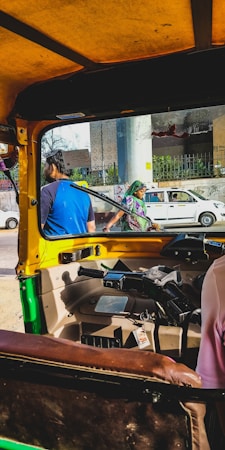 The perspective is from inside an auto rickshaw, with a view of the steering and interior details. Outside, two people are visible&mdash;one wearing a blue shirt and another dressed in traditional clothing with a green headscarf. A white car and a cityscape with an overpass and building structures complete the background.