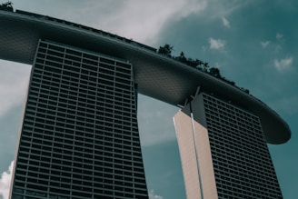 A modern architectural structure with two tall towers connected by a large, curved platform at the top. The platform features greenery, suggesting a garden or park setting. The sky is overcast, giving a moody ambiance to the scene.