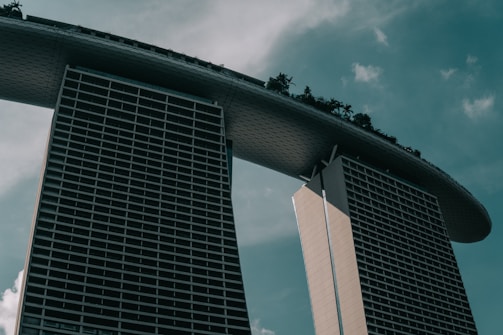 A modern architectural structure with two tall towers connected by a large, curved platform at the top. The platform features greenery, suggesting a garden or park setting. The sky is overcast, giving a moody ambiance to the scene.