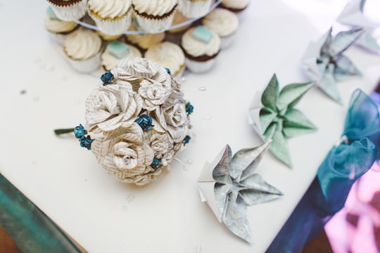 A table adorned with intricately crafted paper flowers made from printed text, accompanied by teal and silver paper origami pieces and a tiered display of cupcakes with white frosting.