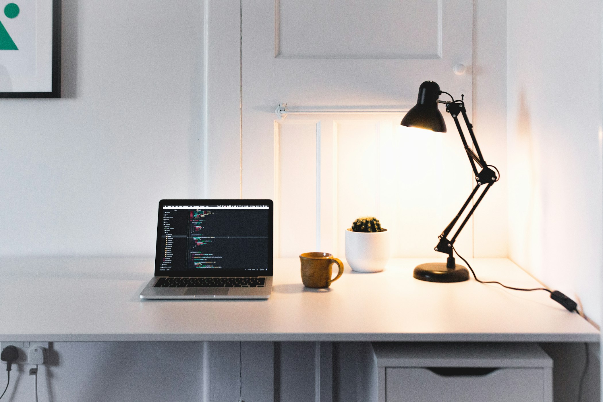 Laptop on top of a white desk, with code on the screen, next to an orange mug, a cactus and a black lamp.