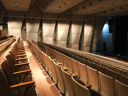 An empty auditorium with multiple rows of beige and brown seats, featuring a modern architectural design with concrete walls and a stage area. The lighting is soft and warm, illuminating the space and creating a quiet, serene atmosphere.