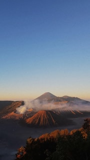 A picturesque landscape of volcanic mountains under a clear blue sky during sunset. The volcanoes are emitting gentle smoke, and the sunlight casts warm shades of orange and brown on the rugged terrain. Lush greenery is visible in the foreground.