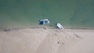 An aerial view of a sandy beach with a small boat docked on the shoreline. A kitesurfing kite is laid out nearby, with some people gathered around it. The sea is calm, and the sand shows patterns created by the wind or water.