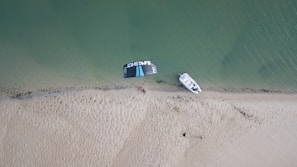An aerial view of a sandy beach with a small boat docked on the shoreline. A kitesurfing kite is laid out nearby, with some people gathered around it. The sea is calm, and the sand shows patterns created by the wind or water.