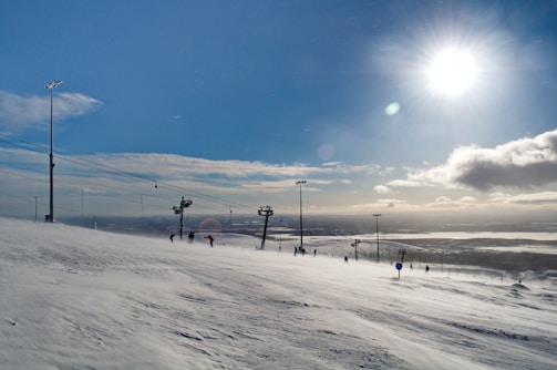 A snowy landscape with a ski slope under a bright sun. Several skiers are descending the slope, and a ski lift is visible on the left side. The sky is clear with a few clouds, creating a serene winter scene.
