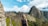 A person with light-colored hair is standing and looking towards the ancient ruins of Machu Picchu. The stone structures of the historical site are set against a backdrop of towering mountains and partly cloudy skies.