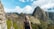 A person with light-colored hair is standing and looking towards the ancient ruins of Machu Picchu. The stone structures of the historical site are set against a backdrop of towering mountains and partly cloudy skies.