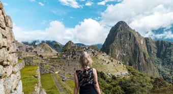 A person with light-colored hair is standing and looking towards the ancient ruins of Machu Picchu. The stone structures of the historical site are set against a backdrop of towering mountains and partly cloudy skies.