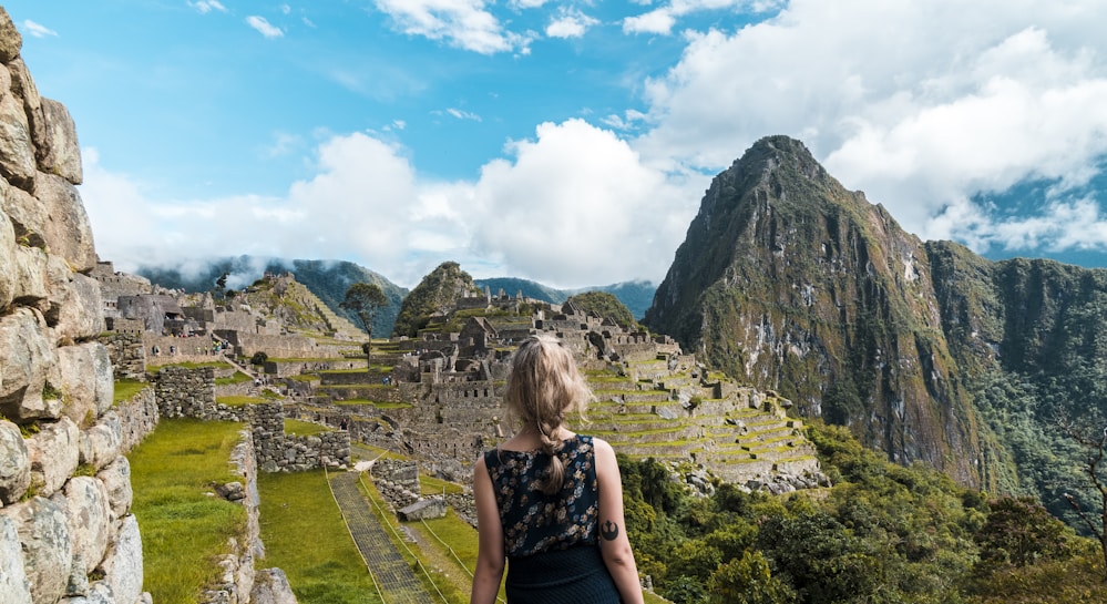 A person with light-colored hair is standing and looking towards the ancient ruins of Machu Picchu. The stone structures of the historical site are set against a backdrop of towering mountains and partly cloudy skies.