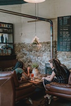 Two people are sitting in a cozy coffee shop with rustic decor, featuring exposed brick walls and a chalkboard menu listing various beverages. They are seated on leather armchairs around a small wooden table that holds drinks and snacks. A potted plant is placed on the table, and soft string lights hang from the ceiling, adding a warm ambiance.