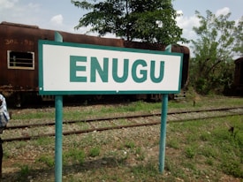 A railway sign with the word 'ENUGU' prominently displayed, set against a backdrop featuring a rusty train car and green foliage. The ground has visible train tracks and scattered greenery.
