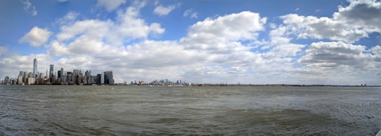 The image features a panoramic view of a city skyline along a waterfront, with numerous skyscrapers including a prominent tall building. The sky is partly cloudy, with soft, fluffy clouds scattered across a blue sky. The water in the foreground appears calm with slight ripples.