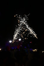 Fireworks burst in a dazzling display against a dark night sky, with several people gathered below. The setting is festive, illuminated by bright, glowing lights and colorful decorations. Hints of red and blue add to the celebratory atmosphere.