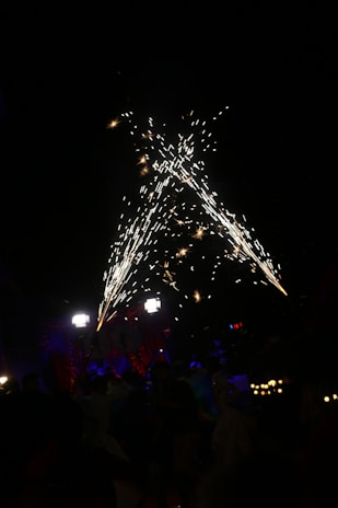 Fireworks burst in a dazzling display against a dark night sky, with several people gathered below. The setting is festive, illuminated by bright, glowing lights and colorful decorations. Hints of red and blue add to the celebratory atmosphere.