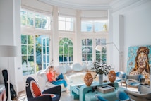 A bright, modern living room with large windows letting in abundant natural light. A person is sitting on a white sofa, using a smartphone. The room features eclectic decor, including a unique black chair with a shark-like design, a light blue coffee table with plants and decorative objects, and a large, colorful portrait painting leaning against the wall. The walls are white, and the overall atmosphere is airy and inviting.